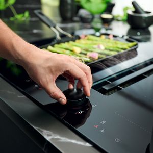 Egy kéz elforgatja a TwistPad® gombot a főzőlapon, miközben a háttérben zöld spárga sül a serpenyőben
