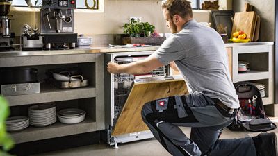 Technician kneels on the floor and installs a dishwasher with a wooden surface in a kitchen.