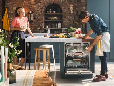 Egy férfi kicsúsztatja a konyhaszigetbe beépített mosogatógép felső kosarát, míg a másik oldalon egy nő várakozik