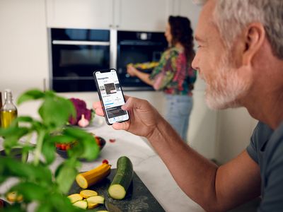 Egy idősebb férfi mobiltelefonjával irányítja a két beépített konyhai készüléket a háttérben, miközben egy nő kezeli a jobb oldalon lévőt