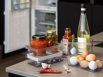 Condiments et œufs dans plusieurs boîtes de rangement Flex Cooling sur le comptoir de la cuisine au premier plan, avec un réfrigérateur et un congélateur ouverts à l'arrière-plan