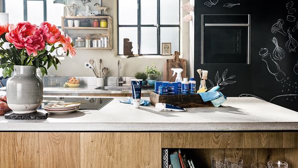 Différents produits de nettoyage et d'entretien et des fleurs roses sur un comptoir de cuisine blanc dans une cuisine aux armoires en bois.