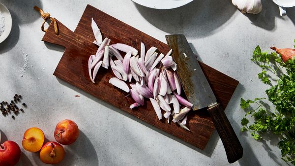 A cutting board with sliced shallots on it beneath a knife.