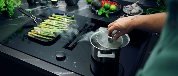 Une main ouvre une casserole pour laisser s'échapper la vapeur dans la hotte intégrée, des asperges vertes cuisent à côté 