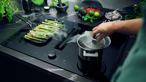 Une main soulève le couvercle d'une casserole pour laisser s'échapper la vapeur dans la hotte intégrée à la table de cuisson, à côté se trouvent des asperges 