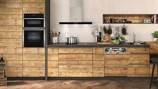 Μεγάλη ξύλινη κουζίνα με κρεμ τοίχους, φούρνους τοποθετημένους τον έναν επάνω στον άλλο και πλήρως εντοιχισμένο πλυντήριο πιάτων ελαφρώς ανοικτό