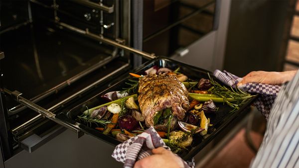 Oven tray filled with meat roast and vegetables being pulled out of oven on extendable rails 