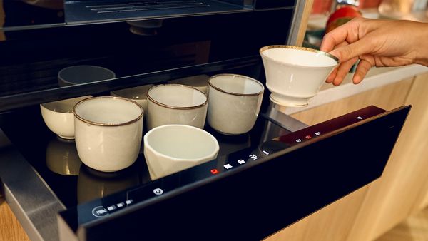 A closer view at an open Warming Drawer beneath an oven, filled with various cups, and a hand is removing a cup