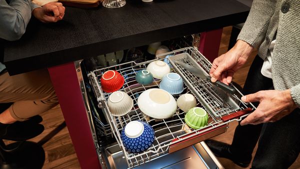 Un lave-vaisselle encastrable en train d'être rempli par une personne, un couteau à droite dans le troisième panier supérieur et de petits bols dans la partie gauche du panier.