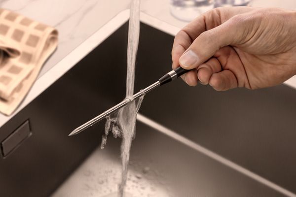 A hand holds a meatprobe under running water in a sink.
