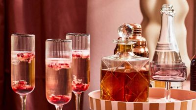 Three champagne flutes with rose petals, a decorative decanter, and sparkling water, set against a pink backdrop.