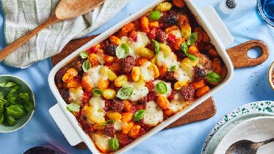 A white baking dish filled gnocchi and sausage in a tomato sauce sits on a light blue tablecloth and is surrounded by other tableware.