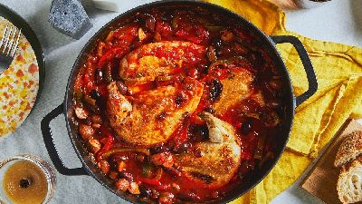 A black pot filled with roasted chicken breasts and a tomato vegetable sauce sits in the middle of a table on a yellow tablecloth and is surrounded by other tableware.