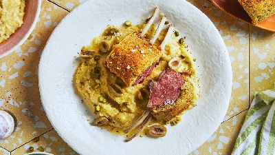 A pistachio crusted rack of lamb sits on polenta on a white plate placed on yellow ceramic tiles and is surrounded by other tableware.