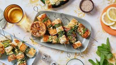 Frittierte Halloumi-Stücke auf Metallspießen liegen auf einem blauen rechteckigen Teller, umgeben von anderen Tellern und Geschirr.