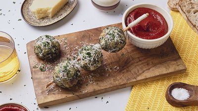 Four spinach and cheese dumplings on a wooden board topped with grated cheese and parsley, served with a bowl of tomato-date sauce and rustic bread on the side.