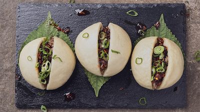 Three steamed pork buns sliced open and filled with spiced ground meat, topped with sliced scallions and chili crisp, served on shiso leaves over a black slate plate.