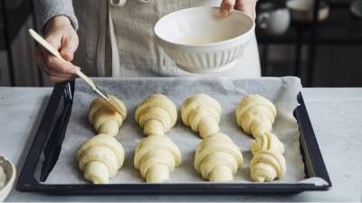 Una persona spazzola il lavaggio delle uova su croissant crudi disposti su una teglia rivestita di carta da forno.
