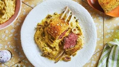 Piatto a base di costolette di agnello impanate su cremosi fagioli schiacciati, guarnite con olive e servite insieme a una fetta di pane alle erbe.