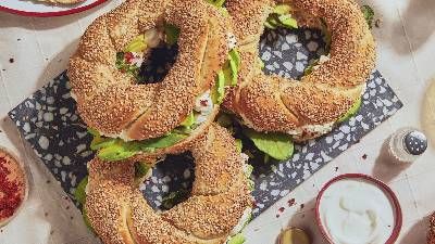 Tre bagel al sesamo ripieni di verdure a foglia verde e condimenti, disposti su un piatto decorativo con contorni di yogurt e spezie.