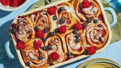 Un piatto al forno di panini alla cannella conditi con glassa e lamponi e mirtilli freschi, apparecchiato su un tavolo colorato. 