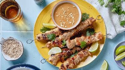 Un piatto giallo vivace contiene spiedini grigliati con erbe aromatiche, spicchi di lime e una ciotola di salsa di arachidi, accompagnati da bevande e guarnizioni.