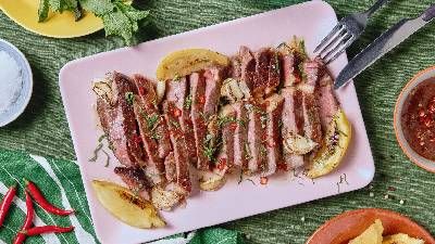 Bistecca grigliata a fette guarnita con erbe aromatiche e peperoncino, servita su un piatto rosa con spicchi di limone e salsa a parte.