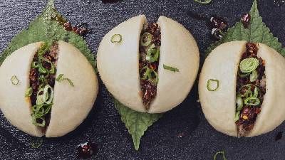 Tre soffici panini bao ripieni di carne saporita e cipollotti, serviti su un piatto di ardesia scura con foglie decorative. 