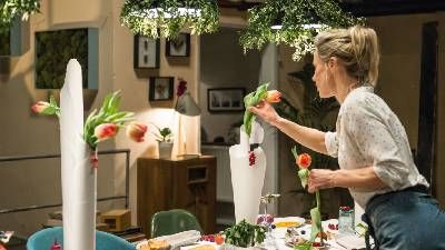 Una donna dispone i fiori su un tavolo da pranzo, circondata da fiori vivaci e verde decorativo in un ambiente accogliente e ben illuminato.