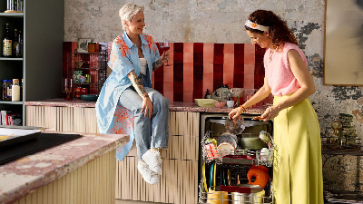 Due donne in cucina, una &egrave; seduta sul piano di lavoro con un bicchiere in mano, mentre l&rsquo;altra sta svuotando la lavastoviglie