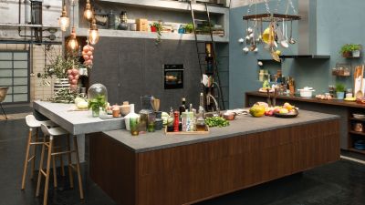 Vista di una cucina open space con isola in legno, piano in cemento e ingredienti freschi disposti sul piano di lavoro