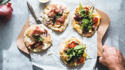 Quattro focacce artigianali con prosciutto, pera, verdure e guarnite con erbe fresche su un tagliere di legno.