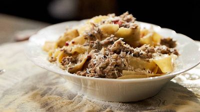 A bowl of pasta with tender meat, topped with grated cheese, sits on a textured tablecloth, inviting and delicious.