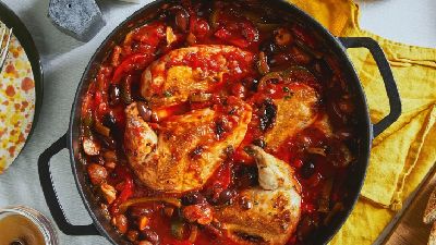 Zwarte braadpan met gestoofde kipstukken in een rijke rode tomatensaus met stukjes paprika en andere groenten; pan staat op een tafel met een geel servet en bijgerechten aan de rand van het beeld.   