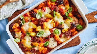 Witte ovenschaal met gebakken gnocchi in tomatensaus, met stukjes chorizo, gesmolten mozzarella en verse basilicumblaadjes; schaal staat op een houten plank met een lichtblauwe doek en een bord aan de rand.   