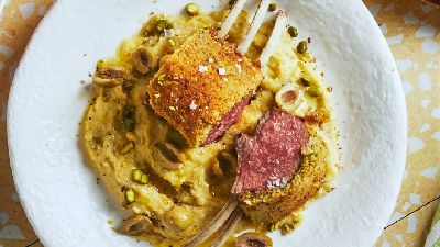 Wit bord met twee krokant gepaneerde lamskoteletten (&eacute;&eacute;n doorgesneden met roze binnenkant) op een romige saus met gesneden olijven en pistachenootjes; een vork steekt in een kotelet, bord op een lichtgeel gevlekt tafelblad.   