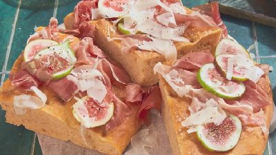 Dikke stukken focaccia op een plank, belegd met dunne plakjes prosciutto, verse vijgen en schaafjes kaas; op een groen betegelde tafel met zoutmolen en extra prosciutto op de achtergrond.   