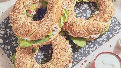 Drie ronde broodjes met sesamzaadjes op een donkere terrazzo-plank, belegd met roomkaas, plakjes avocado en bladgroenten; op een licht tafelkleed staan een kommetje chilivlokken, een kommetje dip/yoghurt en een zoutstrooier naast de plank.   