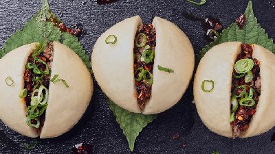 Drie ronde gestoomde bao-broodjes met een donkere, pittige vulling en ringetjes lente-ui, elk geplaatst op een groen blad op een donkere leisteenplaat, met wat chilisausvlekjes eromheen.    