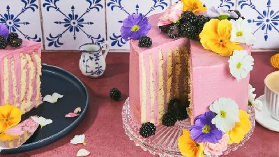 Gelaagde roze taart met een aangesneden stuk, gegarneerd met bramen en kleurrijke bloemen, een stuk op een donker bord met taartspaan, klein melkkanje en een kop koffie voor blauw-witte tegels.   