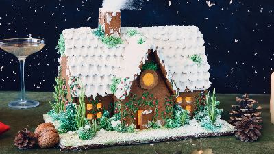 Gemberhuisje met besneeuwde witte glazuurpannen, groene versiering en rode bessen, verlichte ramen, geplaatst op een besneeuwde schaal met dennenappels en walnoten, naast brandende kaarsen en twee coupeglazen tegen een donkere achtergrond.   