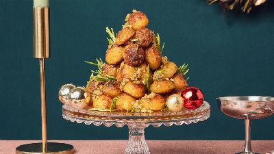 Toren van gekarameliseerde soesjes op een glazen taartplateau, gegarneerd met rozemarijn en gehakte noten, met glinsterende kerstballen en een kaarsenhouder en coupeglas op een donkergroene achtergrond.   
