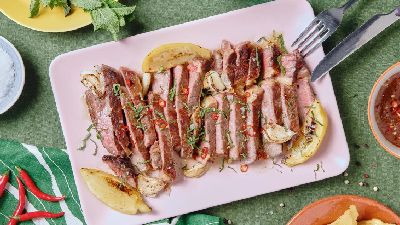 Plakjes medium-rare biefstuk op een roze rechthoekig bord, gegarneerd met gegrilde citroenpartjes, geroosterde knoflook, fijngehakte kruiden en ringetjes rode chili, met een kommetje pittige chilisaus, een schaaltje grof zeezout en verse chilipepers op een groene tafel.   