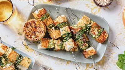 Gegrilde halloumi‑spiesjes met opgerolde courgettelinten, sesamzaad en munt, met gegrilde citroenpartjes op een bloemig tafelkleed.   