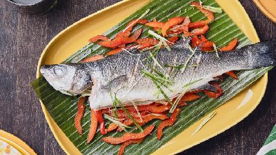 Hele vis op een bananenblad, gegarneerd met reepjes rode paprika en lente-ui, geserveerd op een geel ovaal bord op een donkerhouten tafel, met bestek op een groen servet naast het bord.   