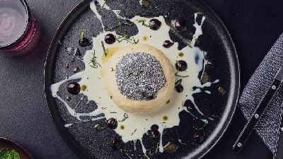 Zwart bord met een rond dessert, bestrooid met poedersuiker en donkere korrels, geserveerd op gespatte witte saus met druppels bessensaus en fijne groene kruiden; bestek op een grijs servet en een glas op tafel.   