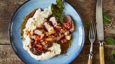 Piatto di agnello affettato con verdure arrostite su pur&egrave; di patate cremoso, guarnito con erbe aromatiche, insieme a coltello e forchetta su un rustico tavolo di legno.