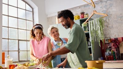 Tre amici sorridenti mentre preparano il cibo insieme in una cucina luminosa e accogliente con ingredienti freschi sul bancone.