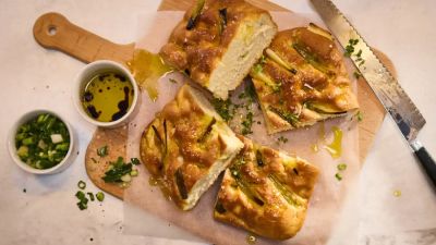 Frisch gebackene Focaccia mit Fr&uuml;hlingszwiebeln, mit Oliven&ouml;l betr&auml;ufelt, serviert auf einem Holzschneidebrett mit Dips.