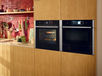 Virtuvėje su mediniu komplektu bei raudonomis plytelėmis šalia viena kitos stovinčios dvi metalinės sidabrinės spalvos orkaitės 